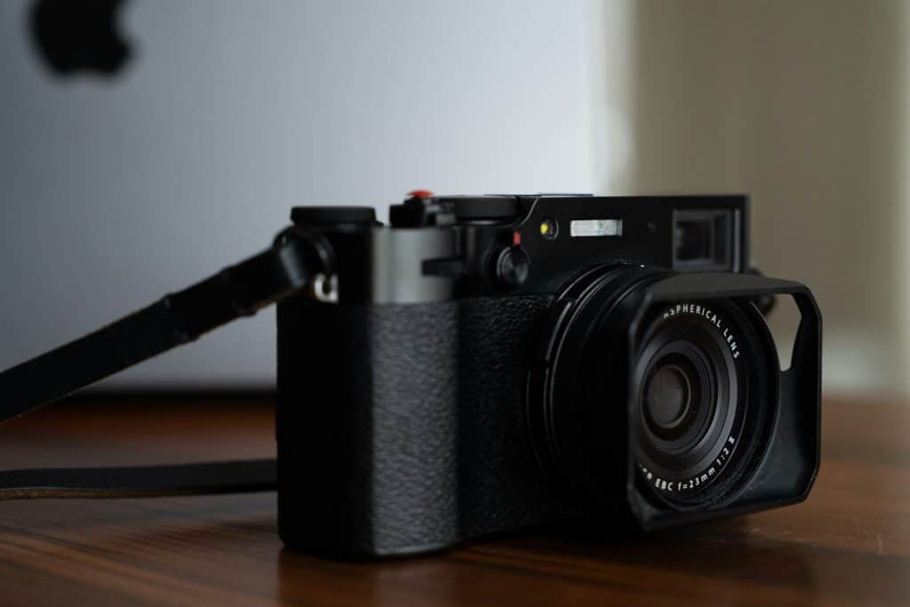 Black Fujifilm x100v mirrorless camera on table with a MacBook Pro in background