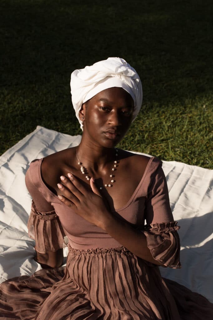 Mujer negra sentada en una manta blanca sobre la hierba, con un vestido de color arcilla y turbante blanco, la mano izquierda apoyada suavemente en el pecho. Foto de Damola Akintunde.