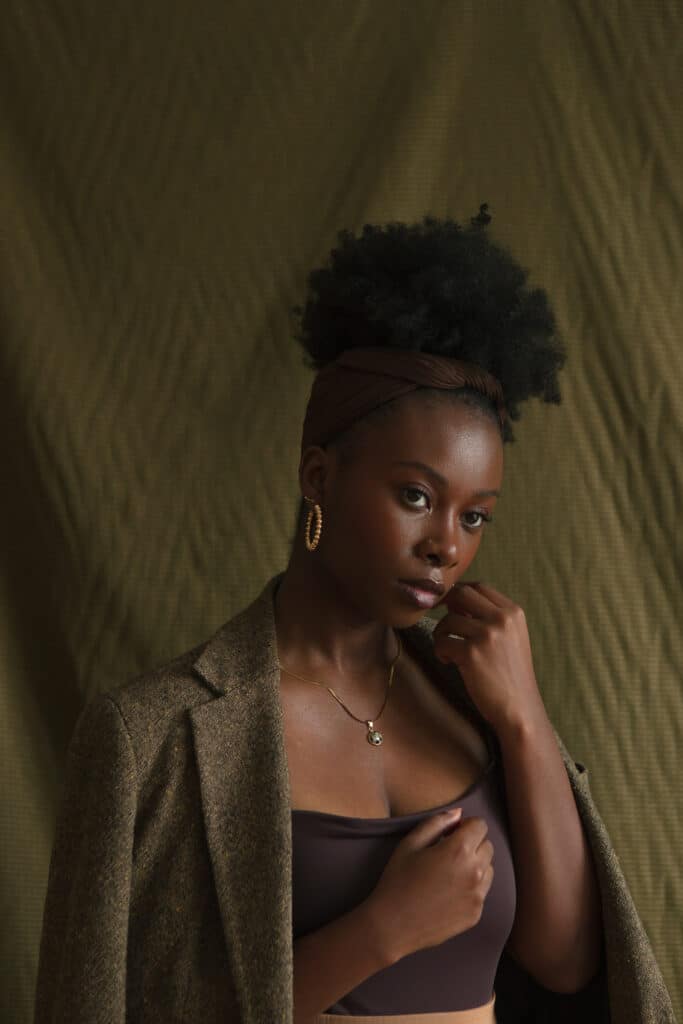 Femme noire posant avec une main fermée tenue doucement près de sa joue, portant un blazer en tweed vert mousse et un haut noir devant une toile de fond verte texturée. Photo prise par Damola Akintunde.