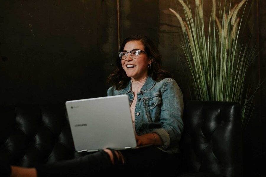 woman wearing glasses and a jean jacket sitting on a black couch with a silver laptop scaled e1702055633145