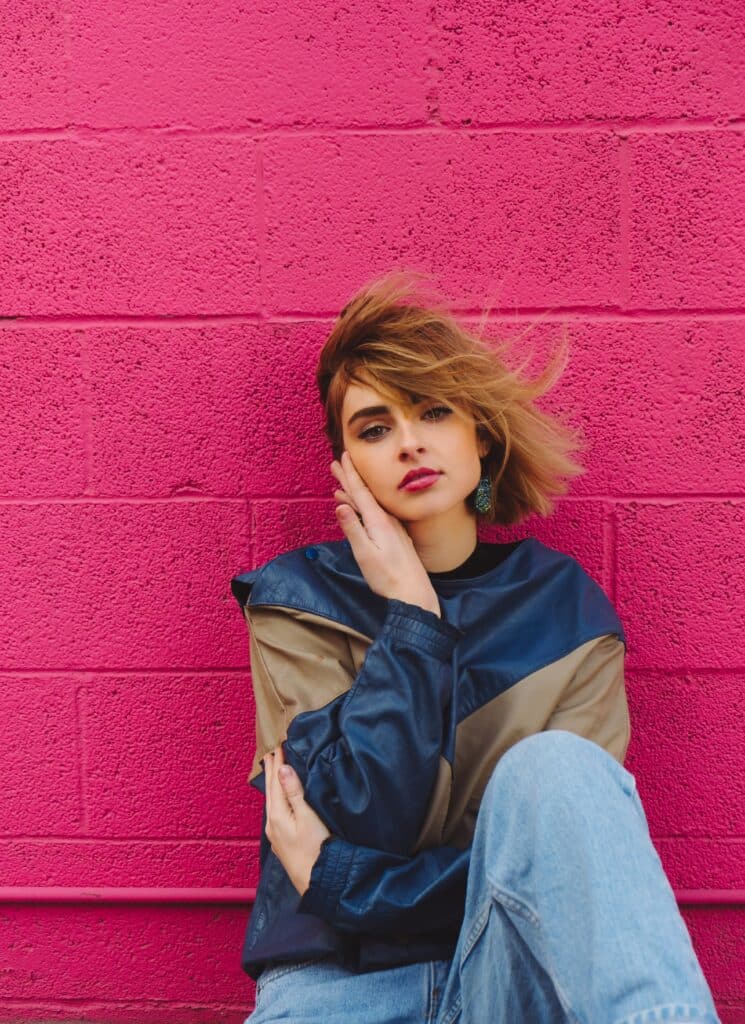 Frau mit marineblauer und taupefarbener Jacke sitzt an einer pinkfarbenen Wand